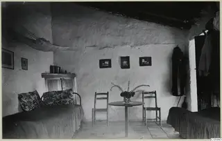 Interior of a whitewashed room at Machairas with two metal beds, a round table, two chairs and framed pictures on the wall.