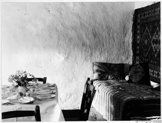 Interior at Machairas with a laid table on the left, a striped bed on the right and a carpet hung on the wall above it.