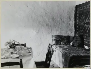 Interior at Machairas with a bed under a hanging carpet and a formally laid table with a white cloth.