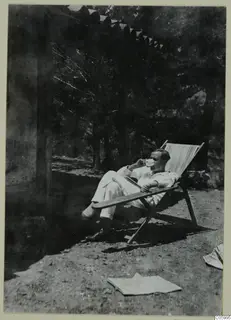 A man in a white suit reclines in a wooden deck chair under trees at Machairas, drinking from a cup.