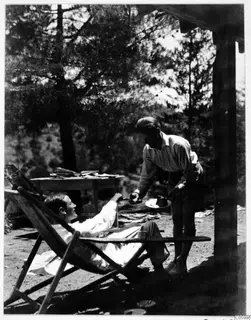 Two men on a terrace at Machairas, one reclining in a deck chair while the other offers him a plate of cake.