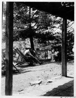 A man and a woman rest in deck chairs on a terrace under a wooden roof, with a large pine tree behind them