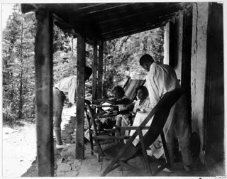 Four people on a shaded terrace of the expedition house at Machairas, two seated at a small table and two standing nearby.