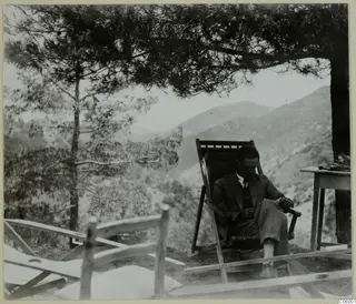 Man seated in a wooden deck chair on a terrace at Machairas, with a pine tree overhead and hills behind.