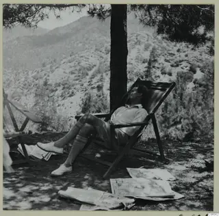 Alfred Westholm resting in a wooden deck chair under a pine tree at Machairas, with newspapers on the ground.