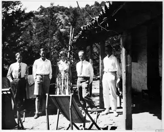Five people stand together on a sandy terrace outside the expedition house at Machairas, with a wooden deckchair in front.