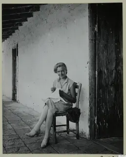 Miss Atterman sitting on a wooden chair on a paved terrace, sewing, beside a whitewashed wall and a wooden door at Machairas.