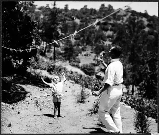 A man with a pipe and a small boy reaching up toward a washing line strung across a dirt track in hilly country