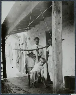 Two boys and two men on a whitewashed terrace; one boy hangs from a pole slung on ropes beneath a beamed roof.