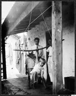Boy hanging from a pole swing under a wooden roof on a paved terrace, with two children and two men nearby.