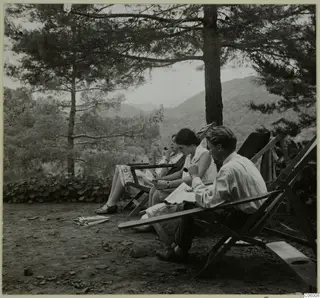 Three people in deck chairs on a terrace under pine trees, reading and smoking a pipe.