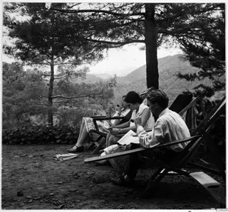 Three people resting in deck chairs on a terrace under pine trees at Machairas, some reading newspapers and smoking.