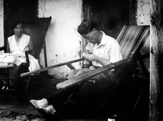 Man darning a sock on a stone terrace at Machairas, with a woman seated in a deck chair beside him
