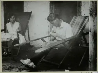 Man darning a sock on a stone terrace, with a woman seated in a deck chair beside him