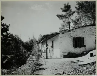 Woman standing on a dirt path beside a rendered stone house with a wooden-posted veranda at Machairas