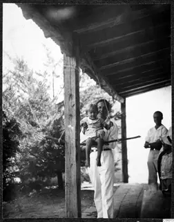 A man holds a small child seated on a swing under a wooden porch roof, with two people standing at the right edge.