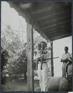 Man in white holds a small boy on a wooden swing bar under a timber terrace roof at Machairas; two men stand at right.