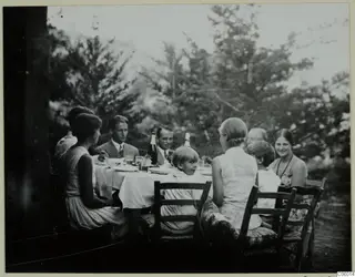 People seated at an outdoor dinner table on the terrace at Machairas, with bottles, glasses and plates under trees.