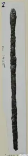 Long, thin, corroded bronze rod with a rounded top, photographed vertically against a pale card mount
