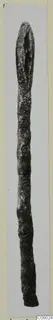 Long slender bronze object, a fragment with a pointed upper end, photographed on a light background.