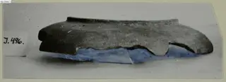 Small find fragment, dark grey with a blue-tinted lower edge, lying on a grey backdrop