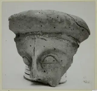 Terracotta face fragment with a flat, cap-like top, a large relief eye and a broken lower face, shown on a tin base.