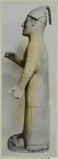 Terracotta standing figure in profile facing left, with a tall cap and incised lines on the robe and arm.