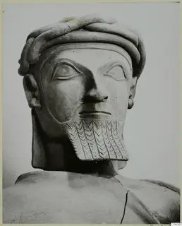 Head and shoulders of a bearded male statue with a twisted headband and a chevron-incised beard