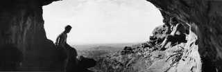 Two people sit at the mouth of a rock cave at Fylleri, Kythrea, looking out over hills and scattered trees.