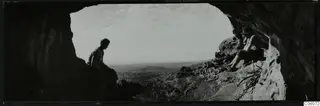 Two people seated at the mouth of a cave at Fylleri, Kythrea, with rolling hills in the distance.