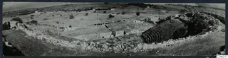 Panoramic view of the theatre at Soli, with stone seating rows curving around a cleared area and a rubble wall in the foreground.