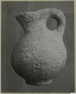 Small stone jug with a rounded body, a spout and a single loop handle, photographed against a grey background