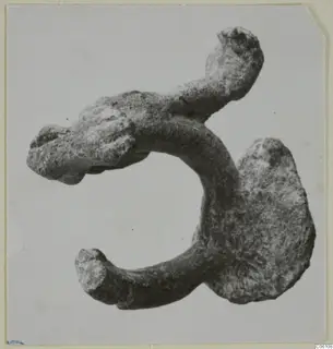Corroded bronze fragment: a thick curved bar with knobbed ends and a broad flattened lobe.