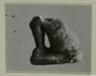 Blurred photograph of a small dark object with a vertical opening, from Ayia Irini, on a cream card mount