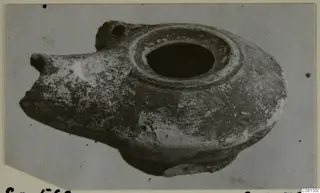 Terracotta oil lamp with a broad filling hole and a pointed spout, photographed on a plain grey backdrop.