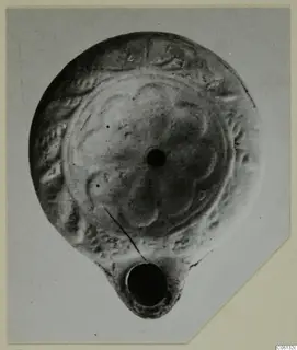 Terracotta oil lamp with a petal-like pattern on its disc, shown on a grey card mount.