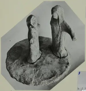 Two terracotta figurines standing on a shared circular base, photographed against a grey backdrop