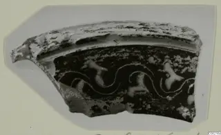 Fragment of a pottery vessel wall with a curved rim, dark surface and pale wavy lines and blobs