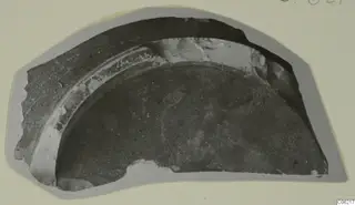 Curved pottery sherd with a dark surface and a pale band along one edge, shown on a grey card mount