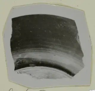 Curved pottery sherd, a fragment with a dark upper surface and a lighter band, shown on a card mount