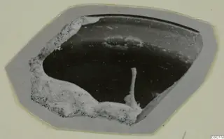 Sherd with a dark, smooth surface and white crusty deposit along one edge, shown on a grey card mount.