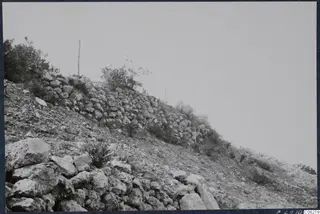 Rough stone wall running up a bare, stony hillside at Vouni, with scrub and a thin pole on the ridge.
