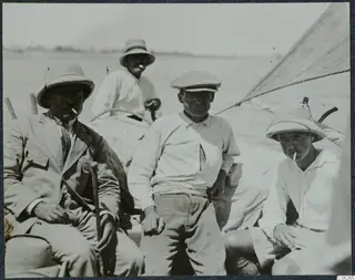 Four men seated in a small sailing boat at Baghazi, with a sail rising at the right edge.