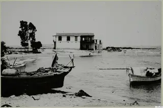 Boats drawn up on a sandy beach, with a two-storey whitewashed building and trees behind them