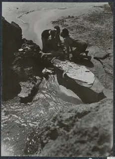 Three people in swimsuits crouched together on a rocky ledge at the water's edge at Kyrenia beach.