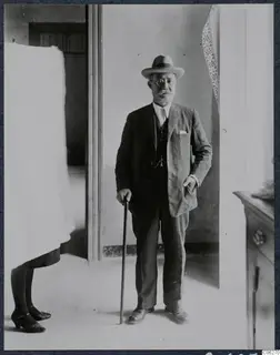 Man in suit, hat and glasses holding a walking stick in a doorway, with part of another person visible at the left edge