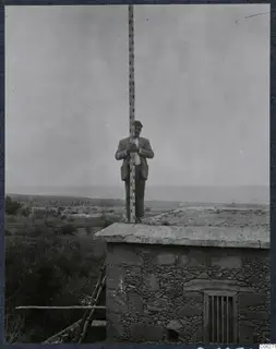 Man holding a measuring rod upright on the flat roof of a stone building at Lapithos