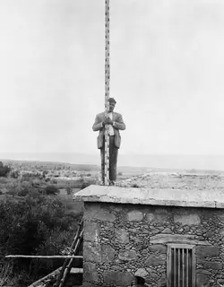 Man holding a tall measuring staff on the flat roof of a stone building at Lapithos