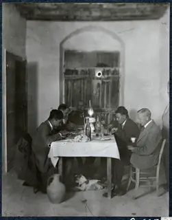 Men eating at a table with a checked cloth in a whitewashed room, with a dog on the floor beneath the table