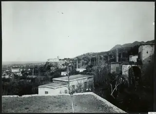 View over the village of Lapithos, with white houses on the slopes, a rocky outcrop with a white building, and mountains beyond.